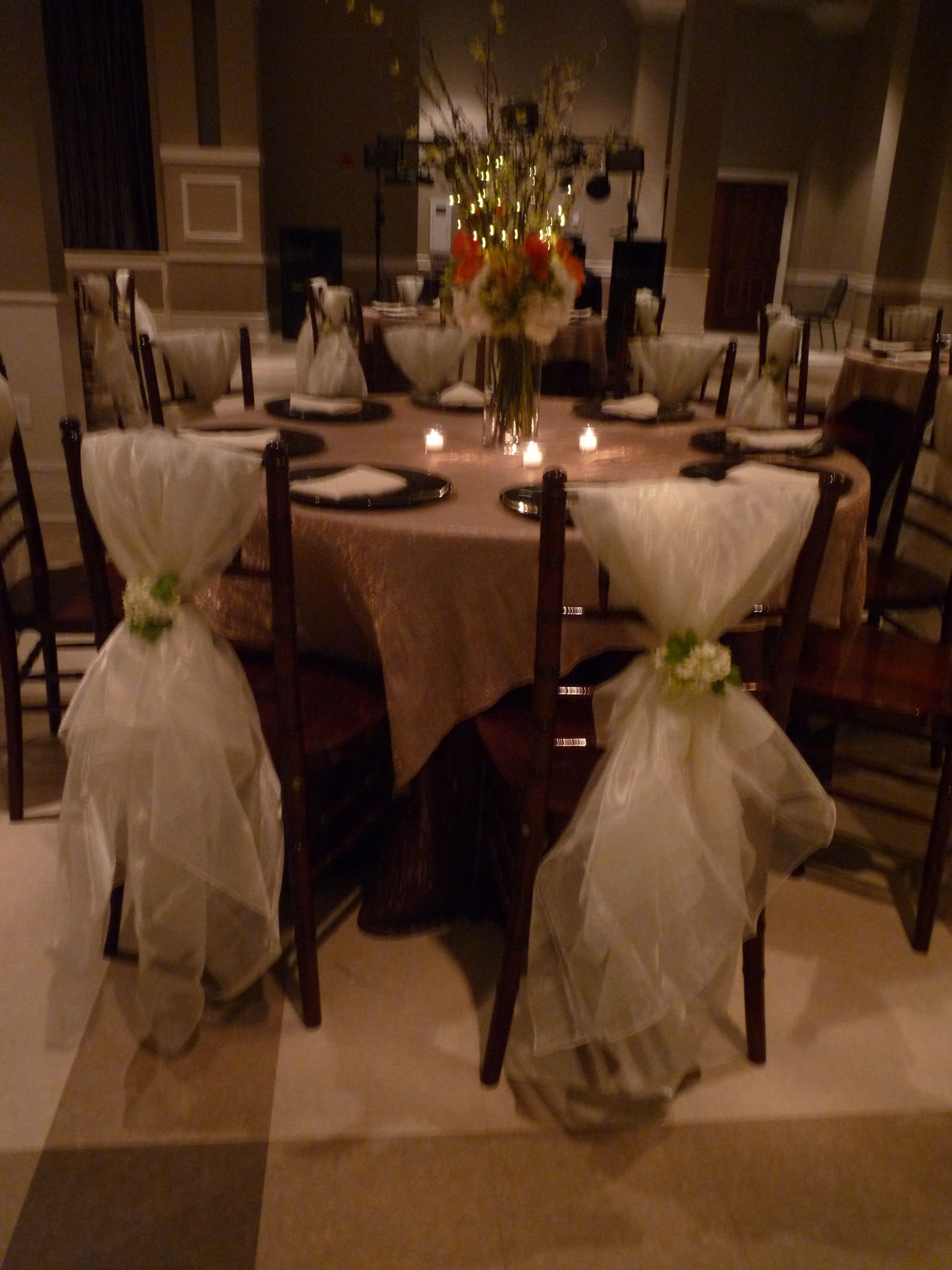 Decorated banquet table with white chair covers, tulle bows, and a floral centerpiece in a dimly lit hall