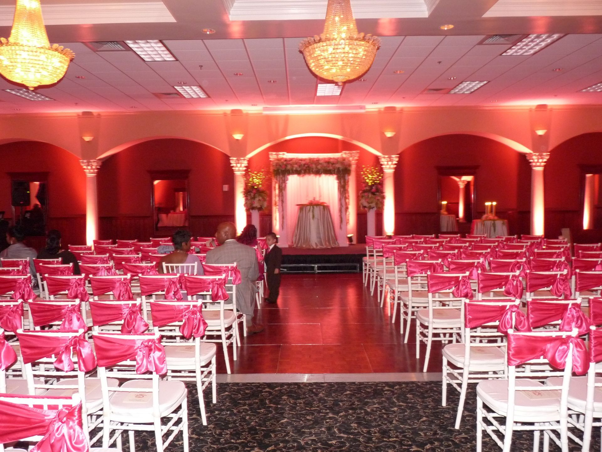 Decorated banquet hall with rows of pink-covered chairs and a red-lit stage at the far end