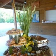 Buffet table with tiered trays of snacks, flowers, and a tall green arrangement in a wooden pavilion