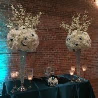 Two white floral topiary arrangements on silver stands beside a black-draped table against a brick wall.