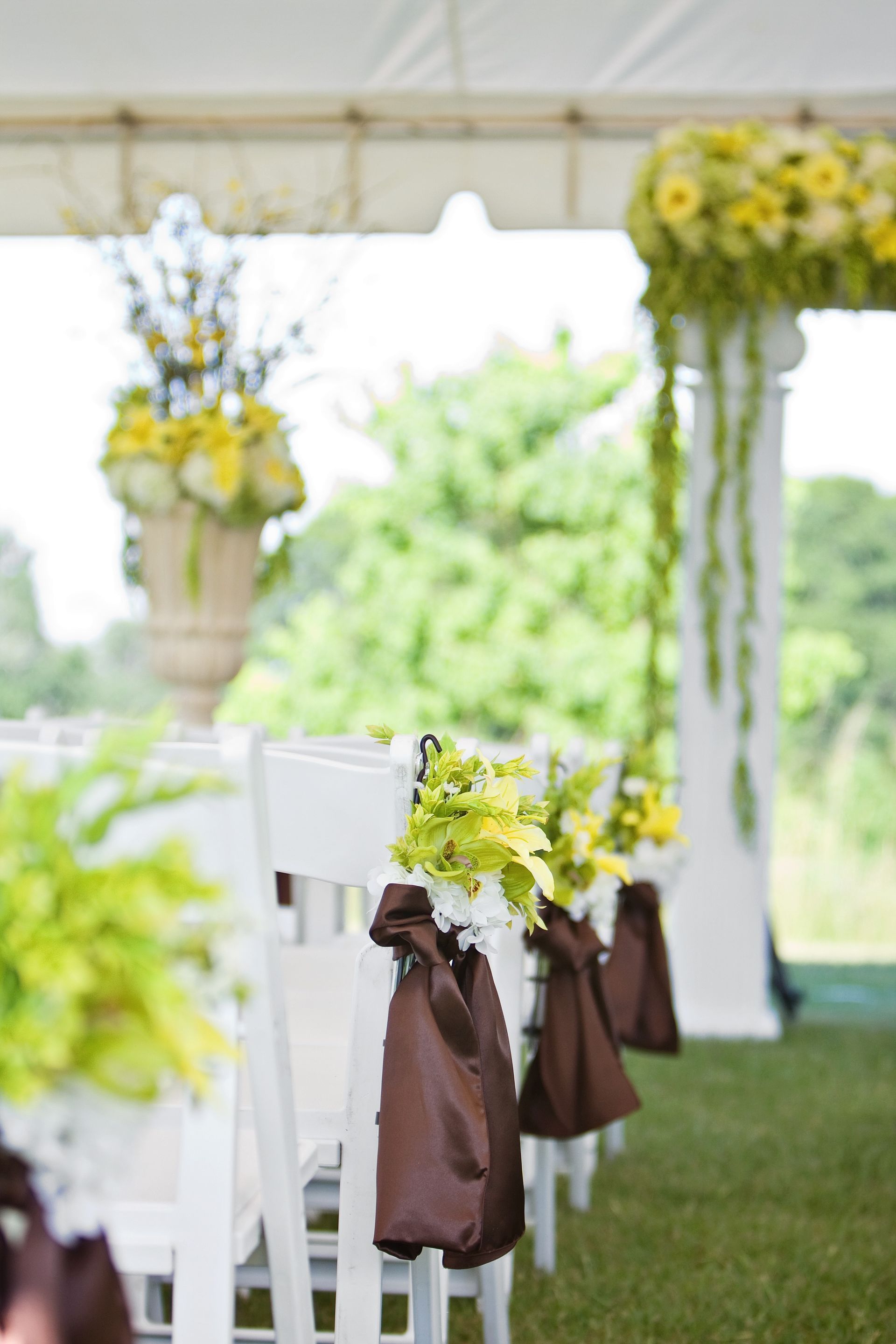 White chairs with brown bows and yellow flower arrangements under a tent at an outdoor event