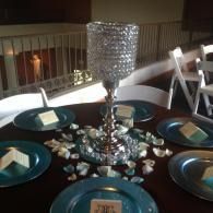 Decorated dining table with teal plates and a silver candle centerpiece, set with scattered crystals.
