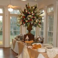 Elegant buffet table with flowers, cakes, and covered dishes in a bright room with large windows