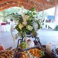 Flower arrangement on a black stand at a decorated outdoor buffet with trays of fruit and desserts.
