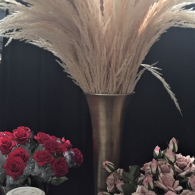 Tall vase with tan pampas grass, flanked by red and pink floral arrangements on a dark backdrop