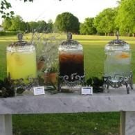 Three glass drink dispensers on a table outdoors: yellow, dark, and clear beverages in a park setting