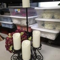 Three white candles on a black holder in front of stacked plastic storage bins and a floral wreath.
