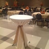 Round cocktail table with white top and beige drape in a banquet hall with seated guests in the background