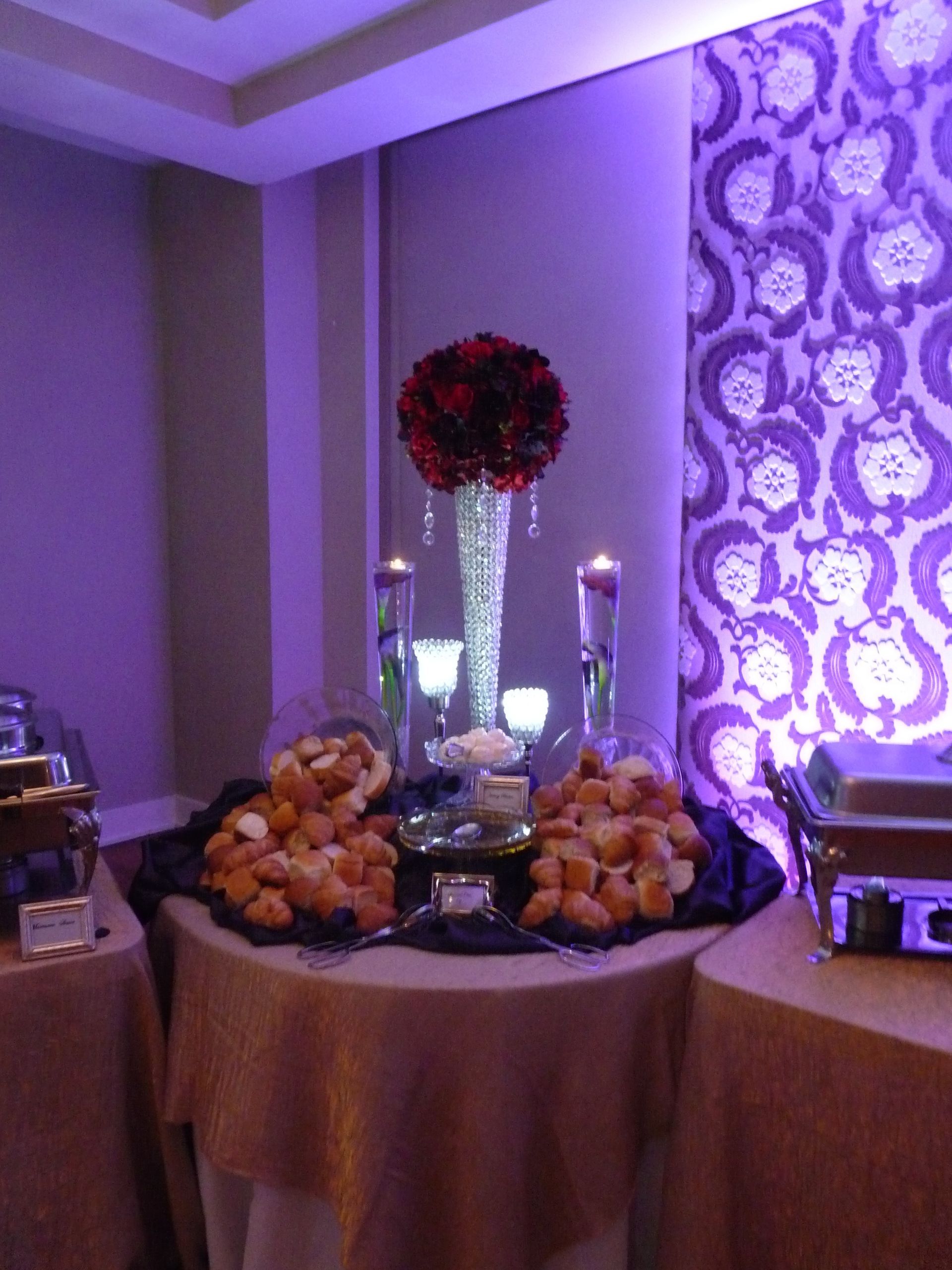 Purple-lit dessert table with pastries, a floral centerpiece, and silver candlesticks at an event venue