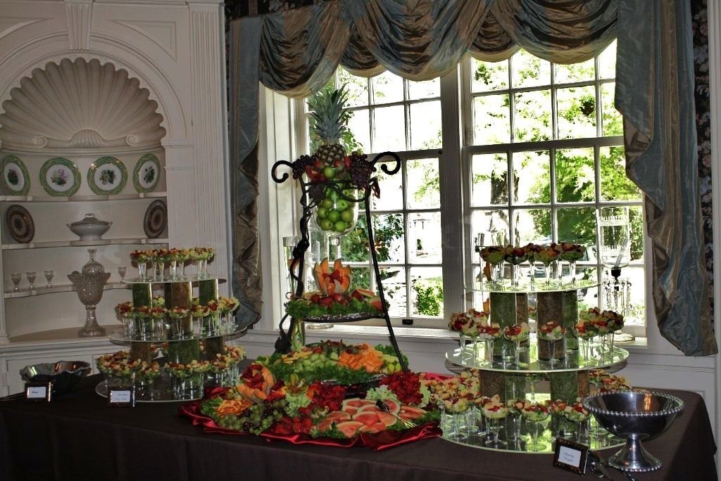 Elegant dessert buffet on a table by a large window, with fruit-topped platters and floral decor