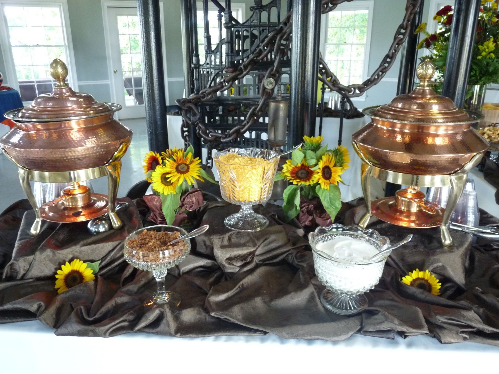 Copper chafing dishes and bowls of food on a black table with sunflowers and dark drapery