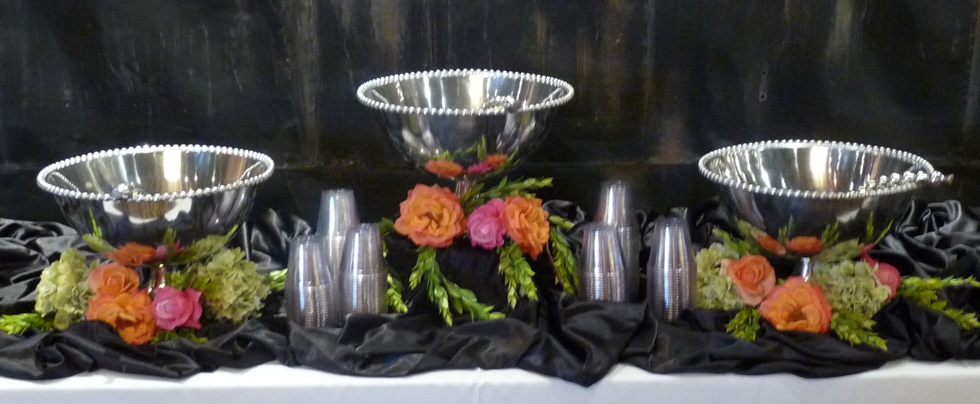 Three silver bowls with orange and pink flowers on a black-draped table with candles