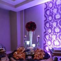 Purple-lit dessert table with bowls of pastries, tall candleholders, and floral centerpiece against patterned wallpaper
