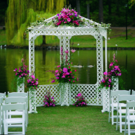 White wedding gazebo with pink flowers by a lake, set on a grassy lawn with white chairs on either side
