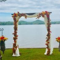 Floral wedding arch by a lake, with grass foreground and distant hills under a cloudy sky