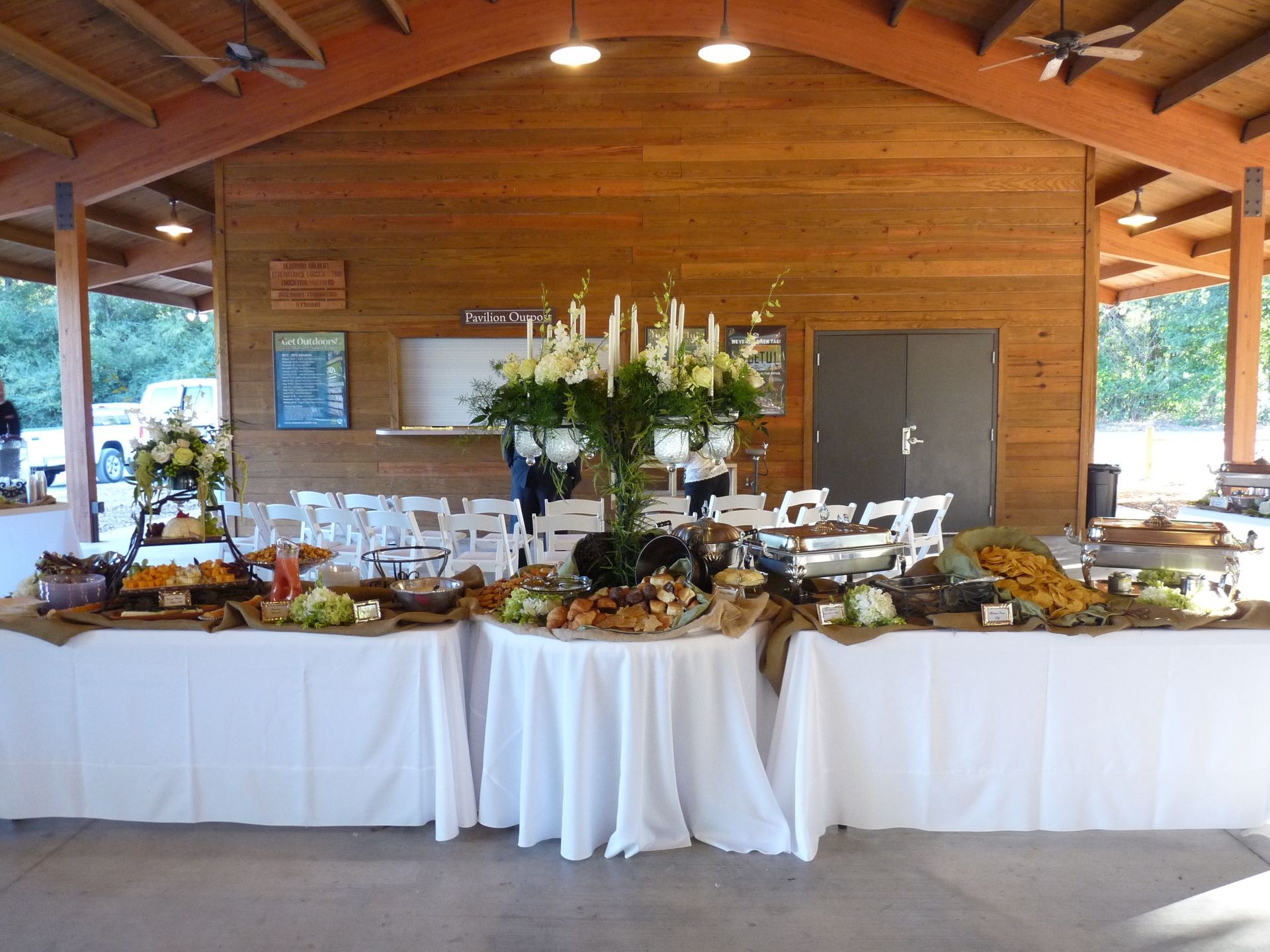 Rustic banquet table with white tablecloths, flowers, and food under a wooden pavilion