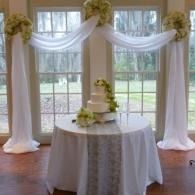 Decorated wedding reception table with white drapes and floral arrangements in front of tall windows