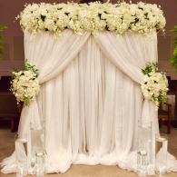 White floral wedding backdrop with draped curtains and hanging flower arrangements, flanked by glass vases