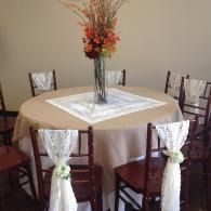 Decorated round dining table with beige cloth, white centerpiece, and four chairs with sheer white sashes