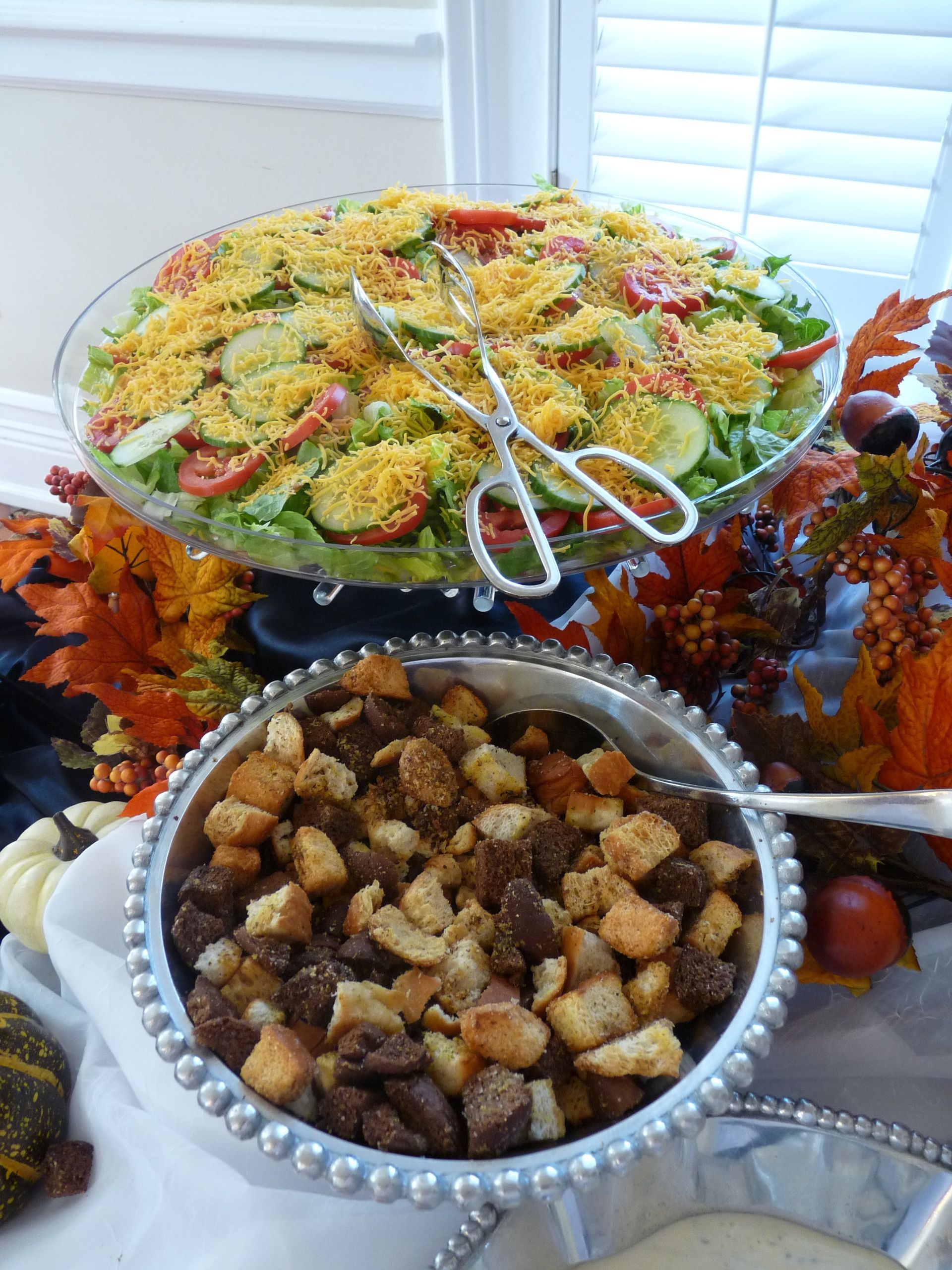 Buffet table with salad, croutons, and autumn leaf decorations, with serving tongs on top