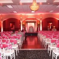 Empty banquet hall with rows of white chairs and pink table settings under red lighting