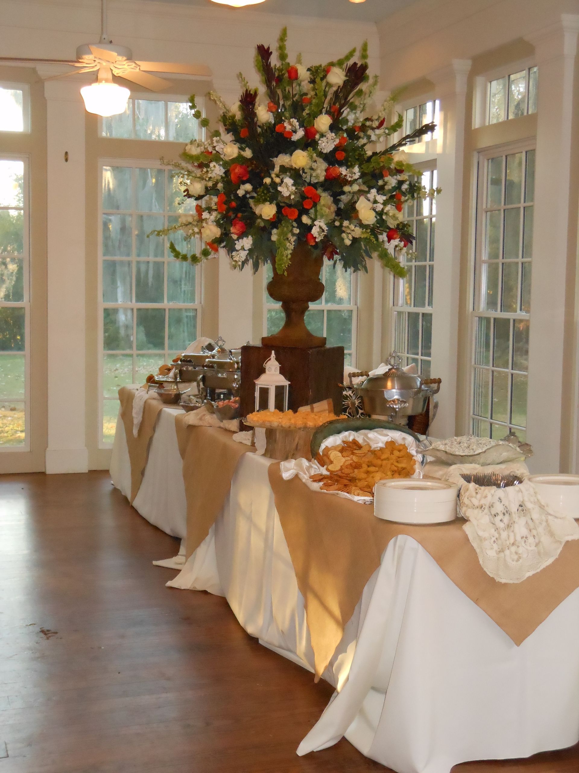 Buffet table with white and gold linens and a tall floral centerpiece in a bright room