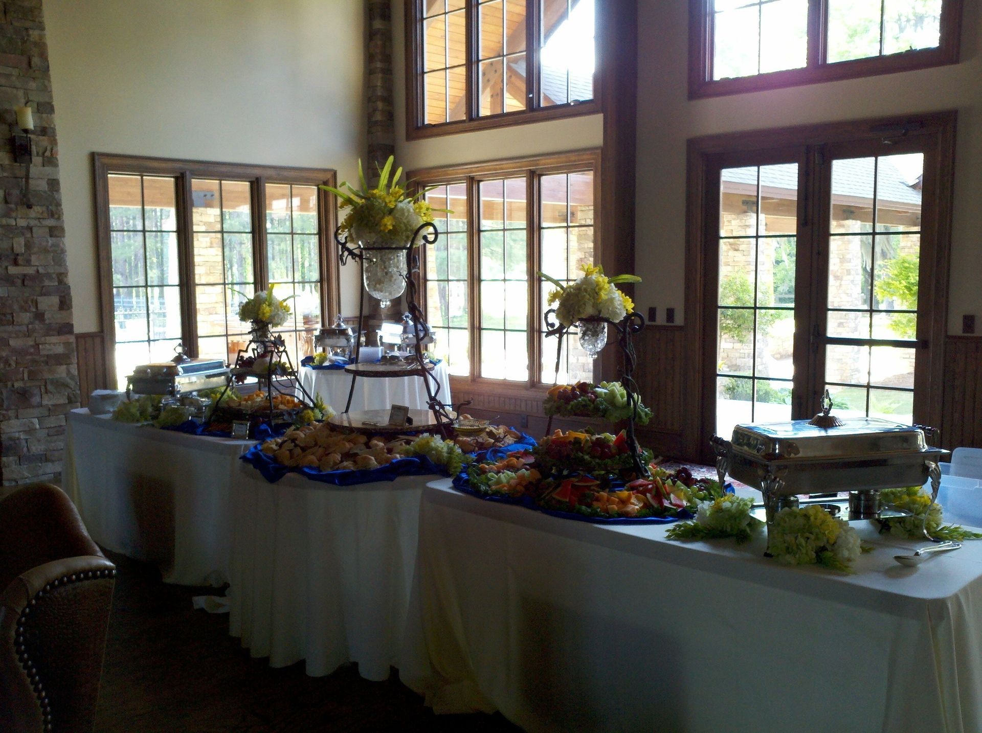 Buffet table with floral arrangements and trays of food in a bright room with large windows