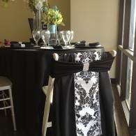 Black cocktail table with black-and-white damask tablecloth and glass vases on top near a window