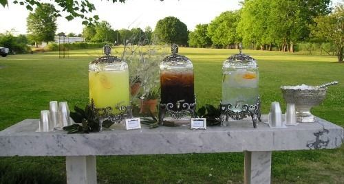 Three drink dispensers on a white table outdoors in a park, with grass and trees in the background