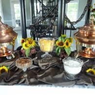 Copper chafing dishes and bowls of fruit and flowers arranged on a dark tablecloth at a buffet display