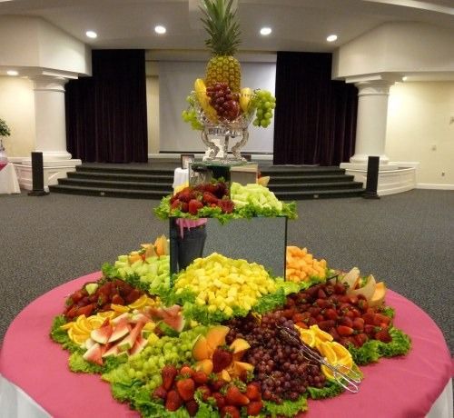 Fruit display on a pink tablecloth in a banquet hall, with a tall pineapple centerpiece and assorted fruits around it