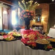 Buffet table with assorted foods and a tall floral centerpiece in a warmly lit brick-walled room