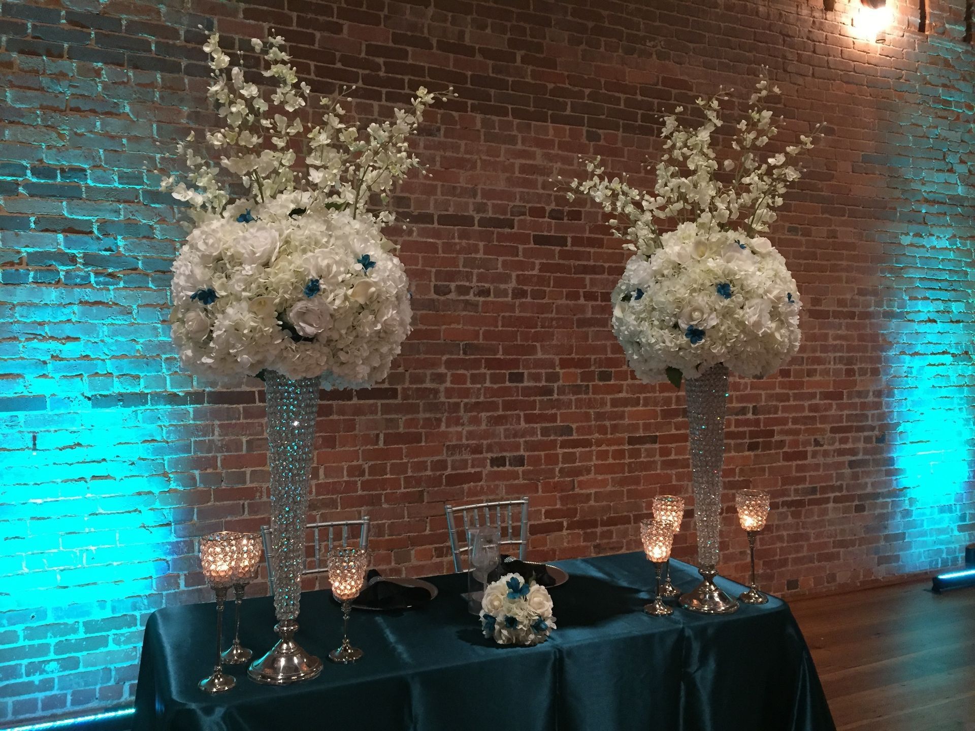 Elegant floral arrangements on tall silver stands beside a teal-draped table against a brick wall.