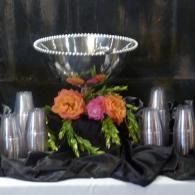 Silver bowl centerpiece with orange and pink flowers, surrounded by stacked metal cups on a black cloth.