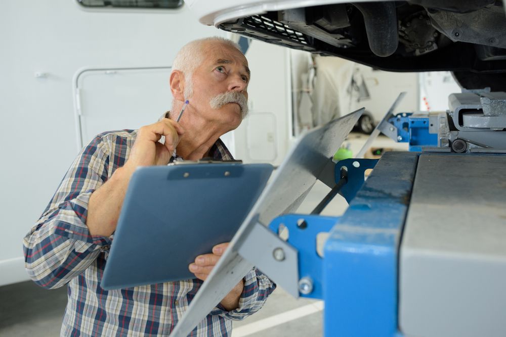 Senior Man Inspecting Vehicle Undercarriage With Clipboard in Auto Repair Shop — Darren's Caravan Repairs in Lake Macquarie, NSW
