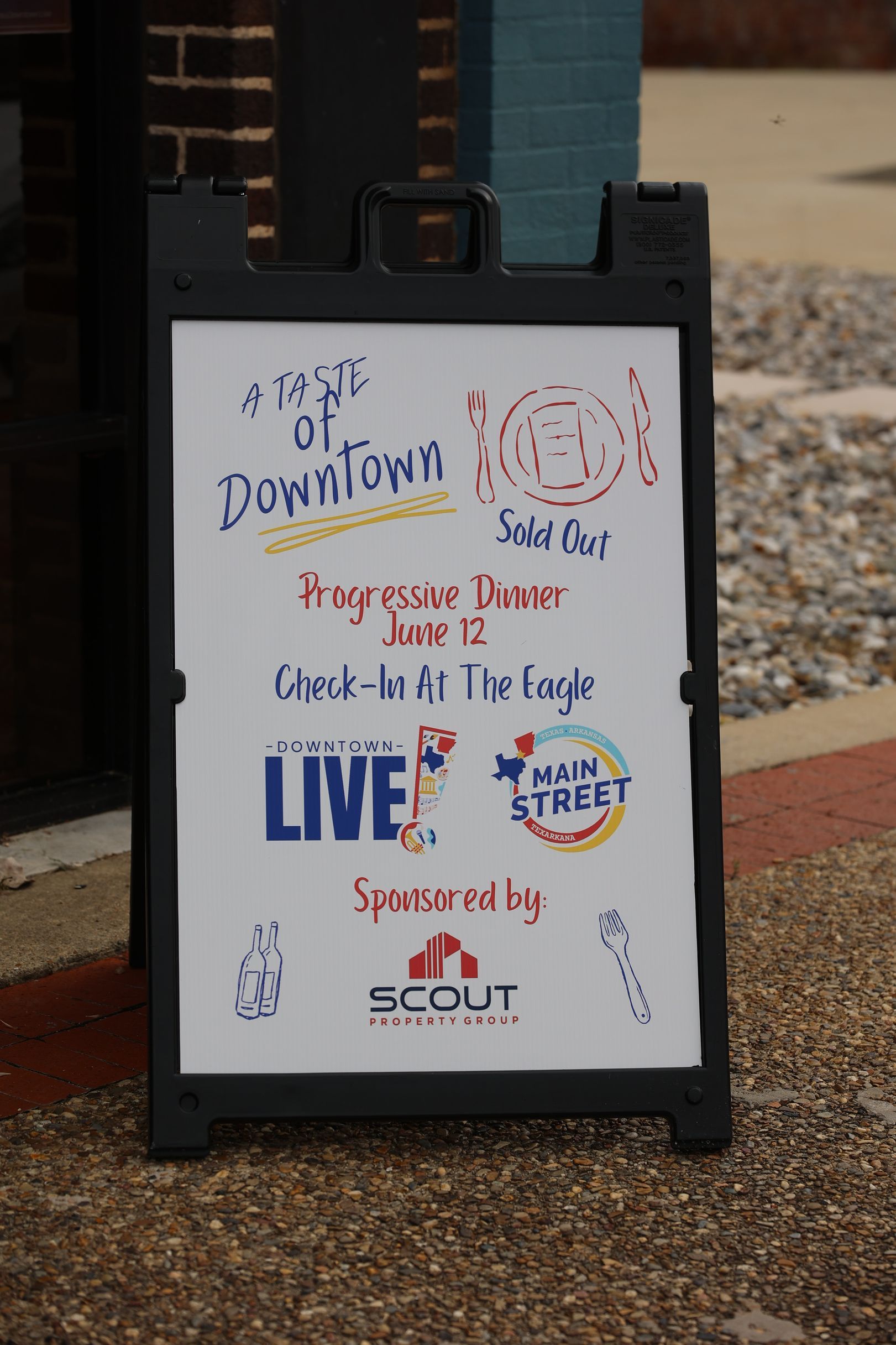 A black sidewalk sign with colorful handwritten messages promoting Downtown LIVE in Texarkana