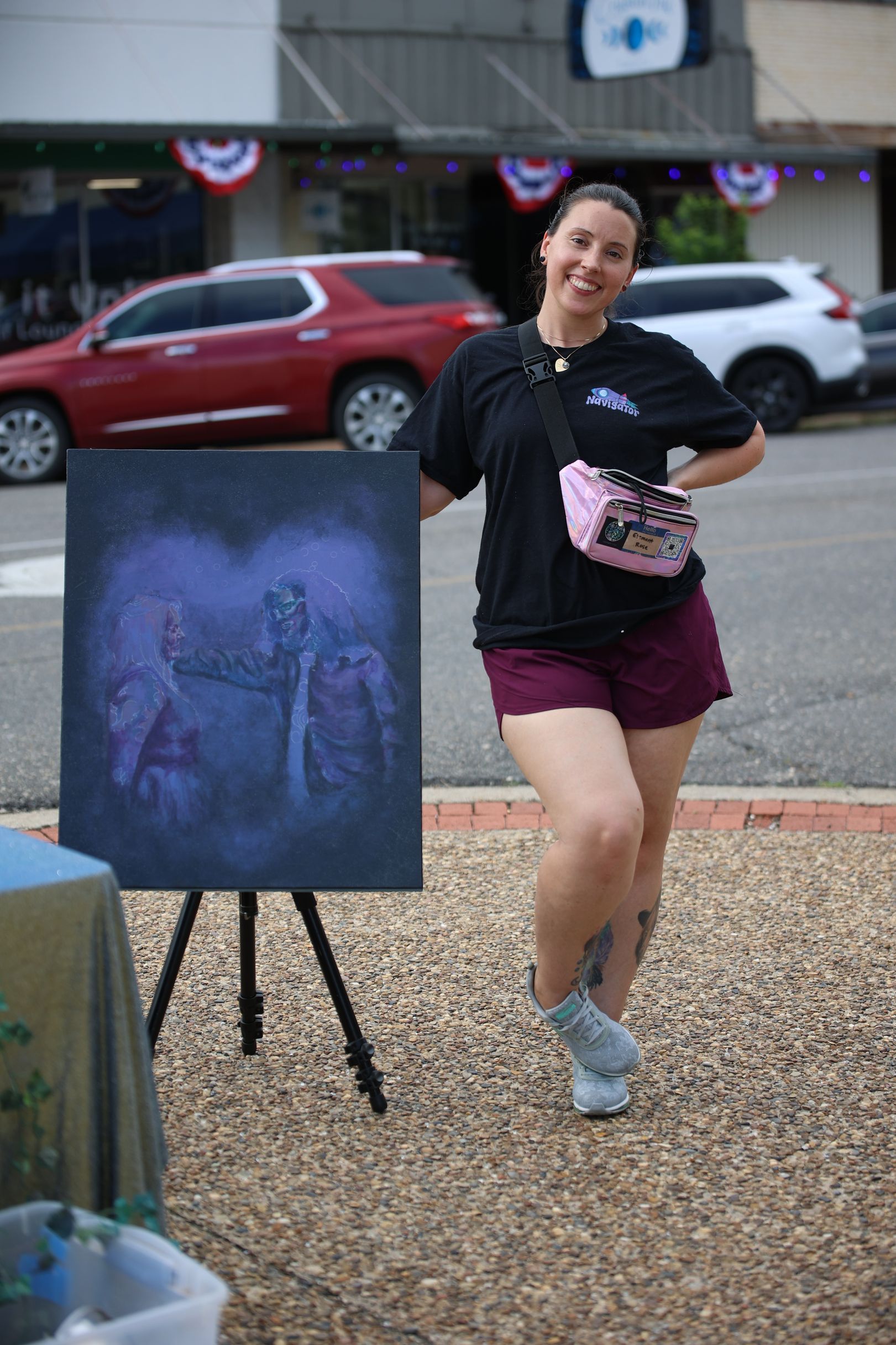 Woman standing beside a painting on an easel outdoors, holding a pink item in front of a building.