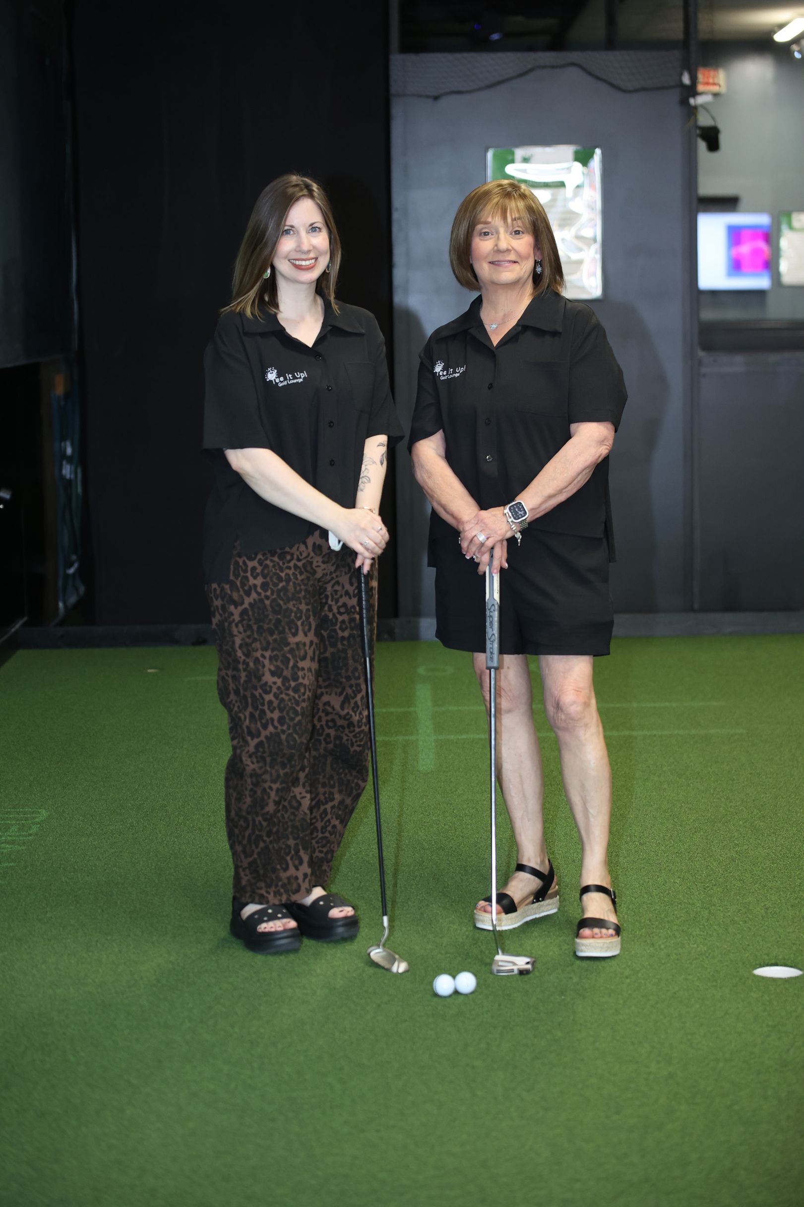 Two women stand on indoor putting green, holding golf clubs beside a hole with golf balls.