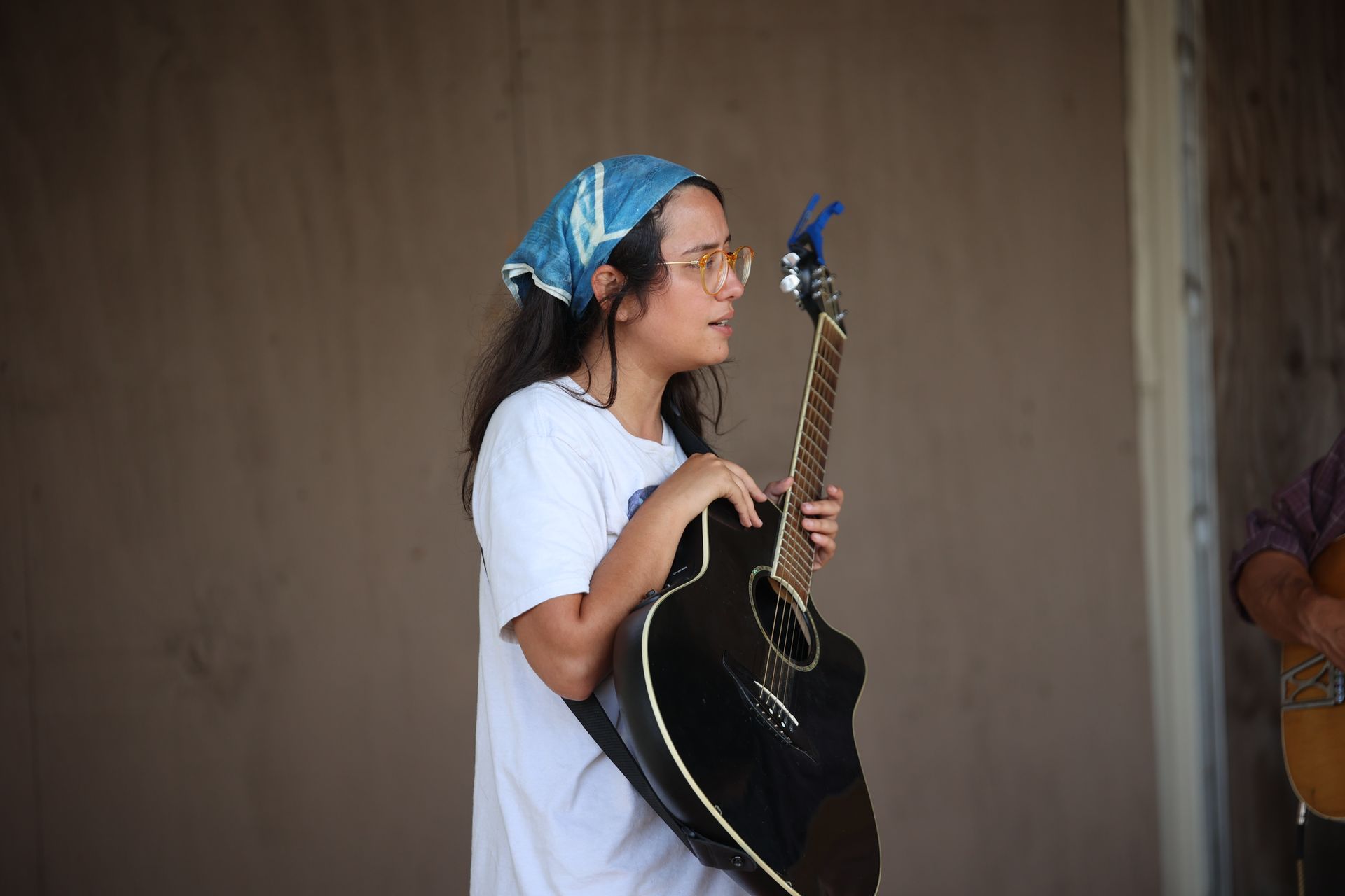 Woman in a blue headscarf holding a black acoustic guitar, standing indoors and looking left