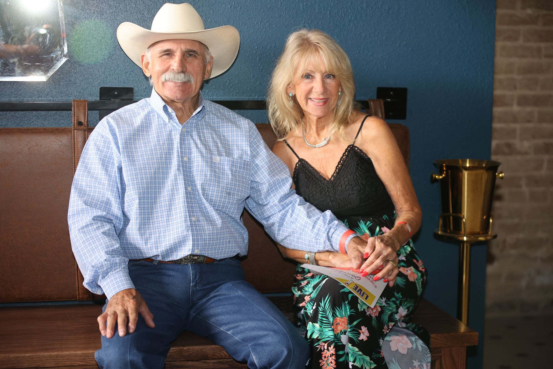 Two people seated indoors, smiling and holding hands; man wears a cowboy hat, woman in a black top and floral skirt.