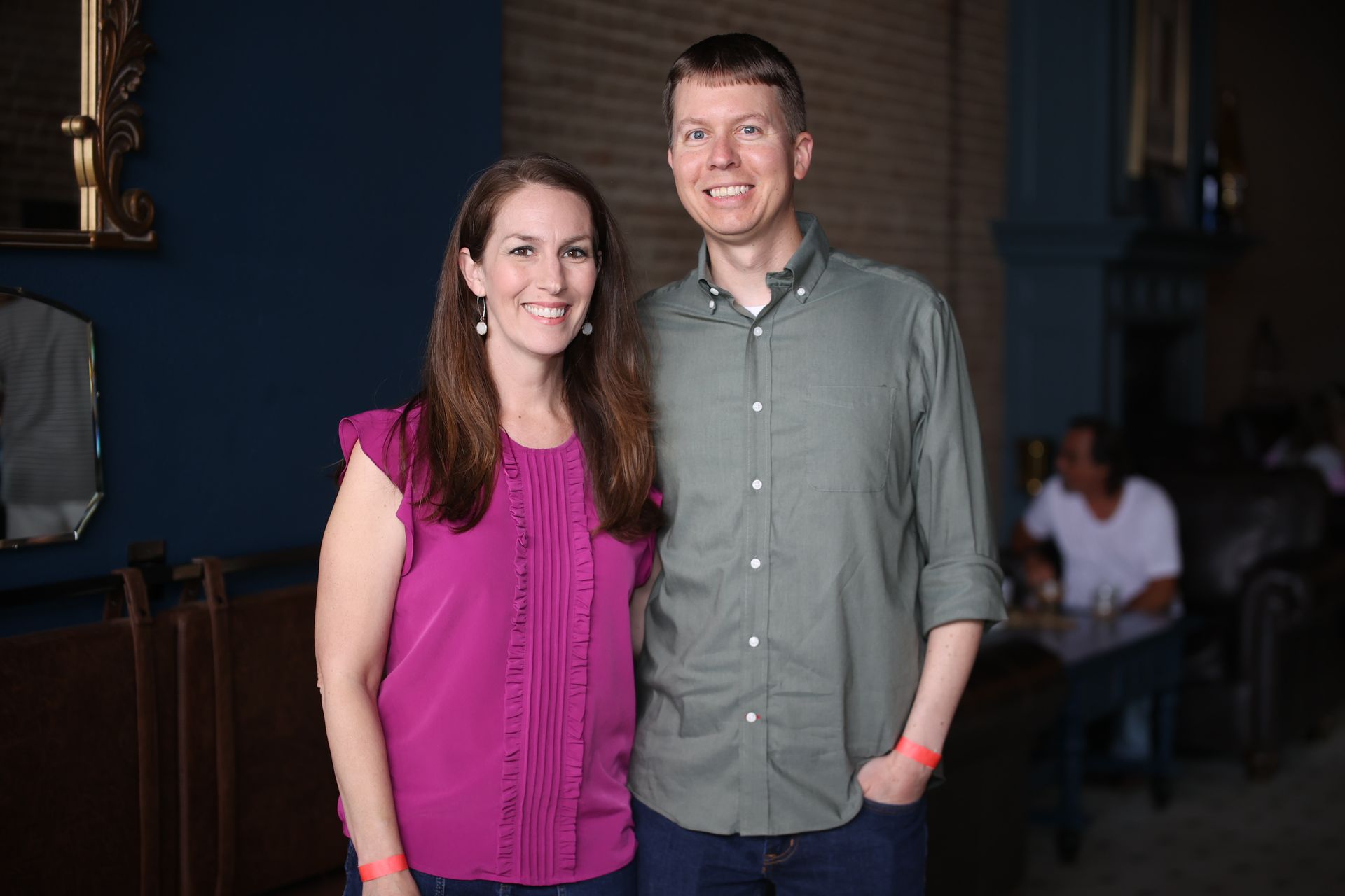 Two people standing indoors, smiling at the camera; woman in a magenta blouse, man in a gray shirt.