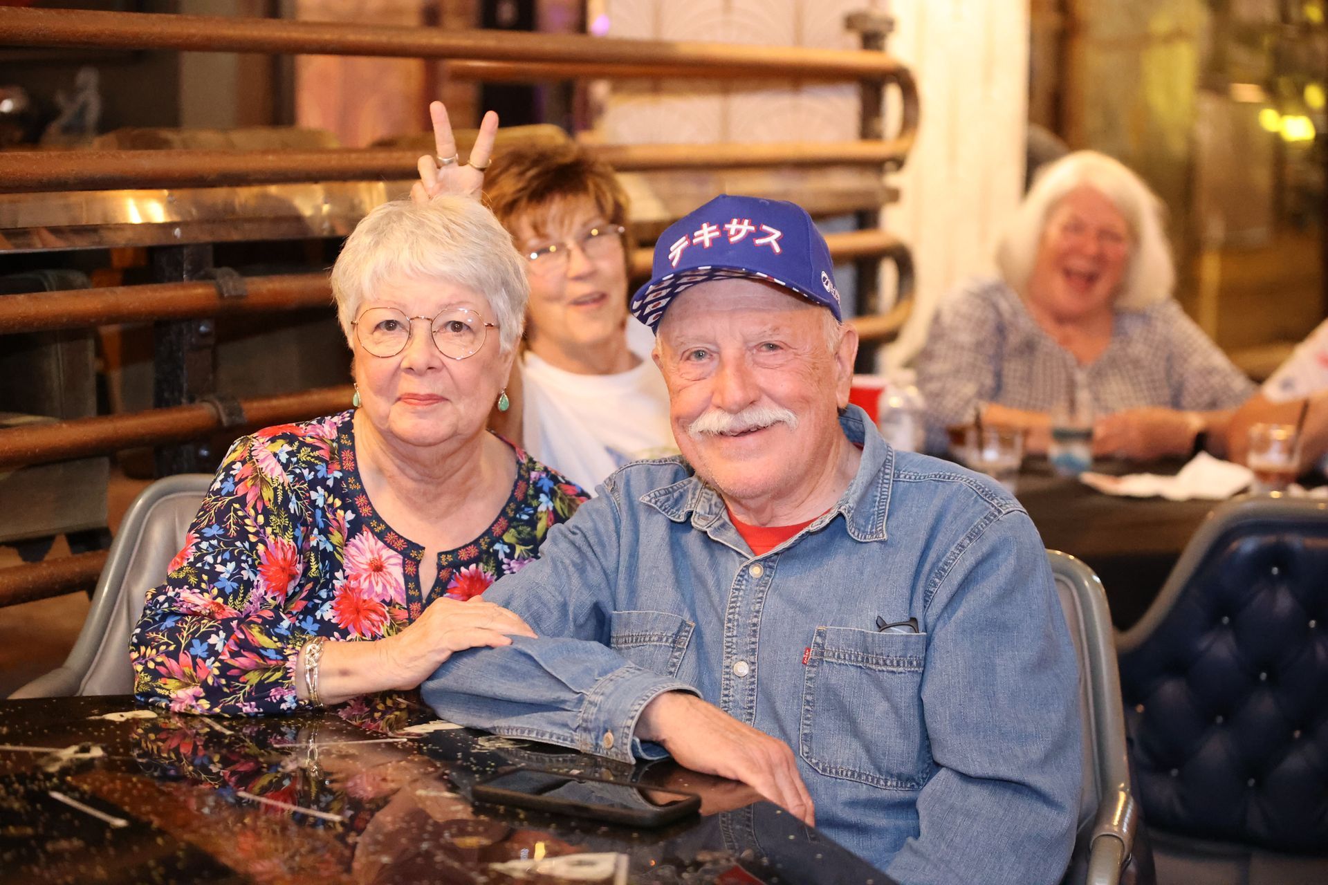 Three people smiling at a restaurant table, with an elderly man in a blue cap in the foreground.