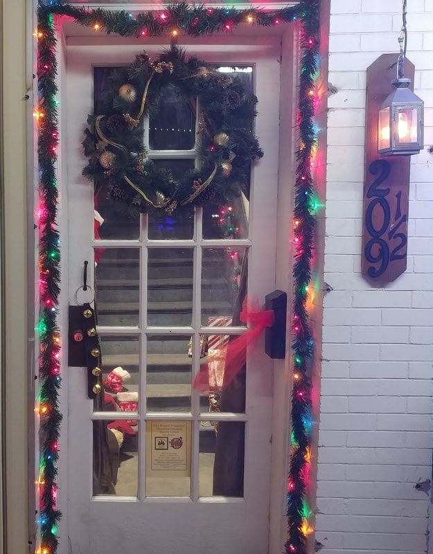 White door decorated with Christmas lights, wreath, and garland. A lantern and a sign with the number 202 are also present.