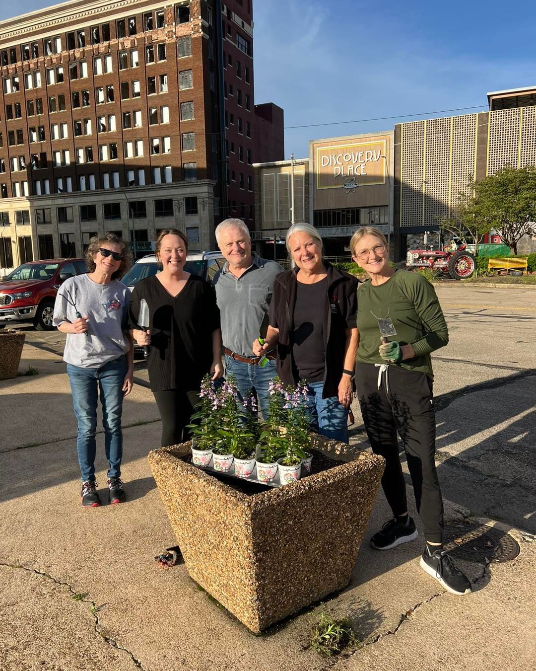 Five people pose with a planter filled with purple flowers, in front of a city building.