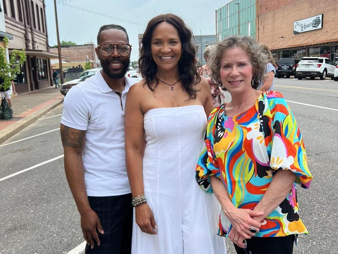 Three people smiling on a street; a man in white shirt, woman in white dress, woman in colorful top.