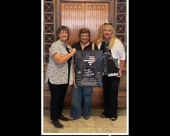 Three women holding a framed poster in front of an ornate wooden door.