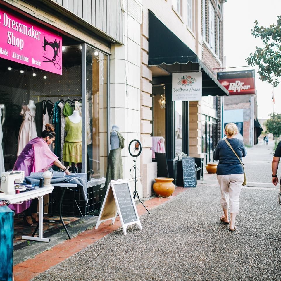 Street scene with a seamstress working in a shop window, two people walking on sidewalk.