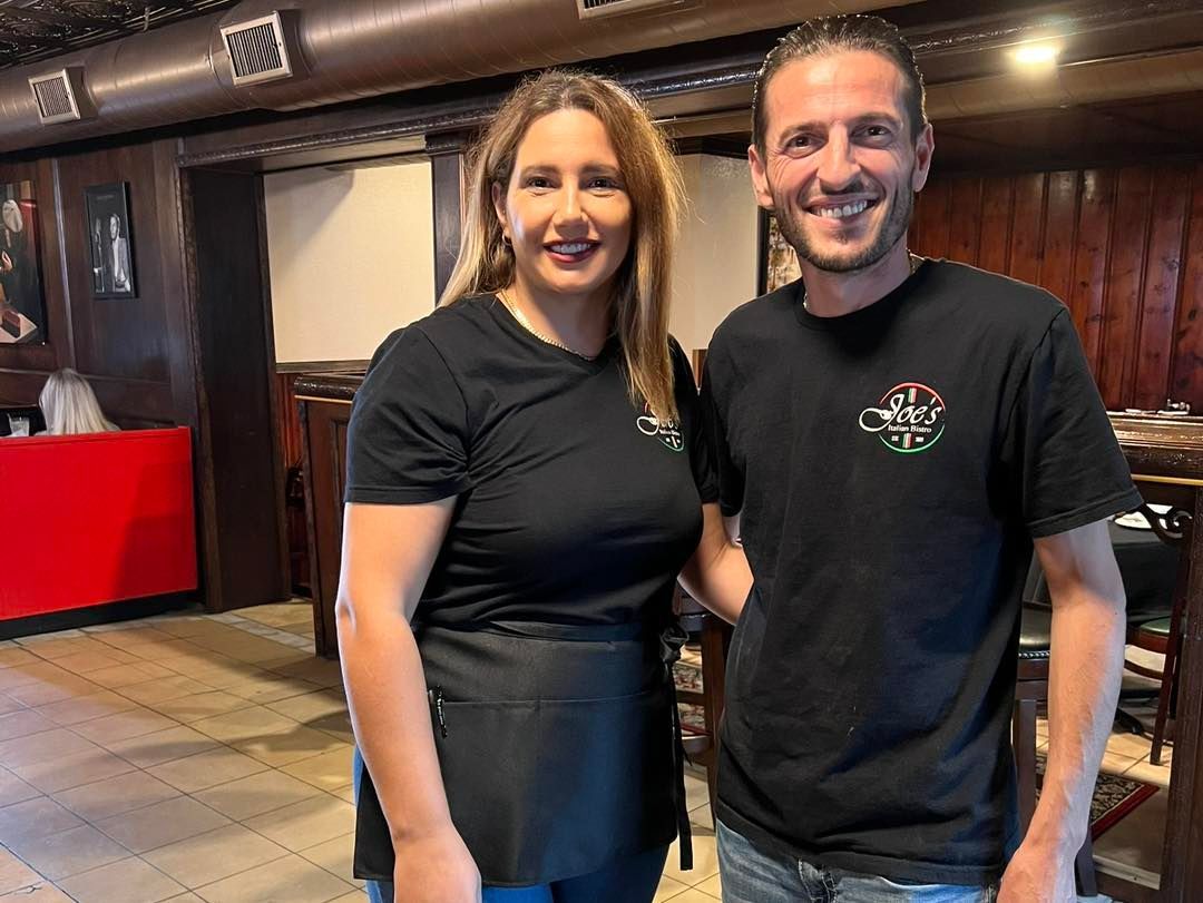 Two people, a woman and a man, smiling, wearing black shirts, inside a restaurant.