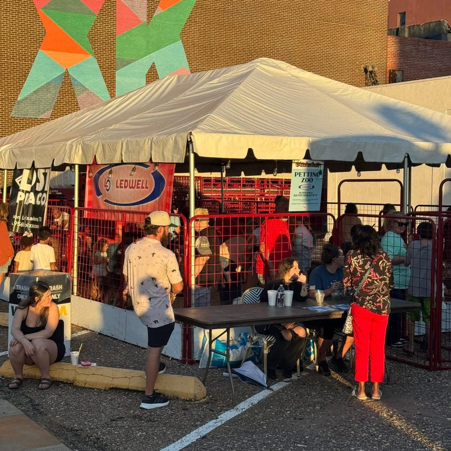 Outdoor event: People under a tent, red fencing, and colorful mural background.