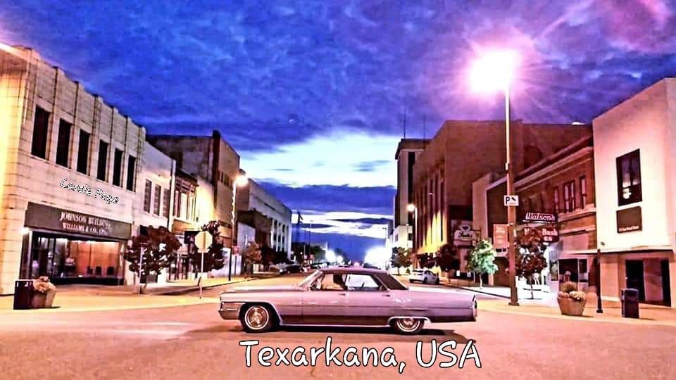 Classic car driving down a street at dusk in downtown Texarkana, USA. Buildings line the street, illuminated by streetlights.